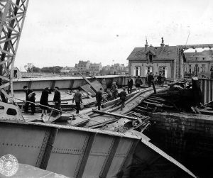 Civilians cross a wrecked bridge in Cherbourg, 1944 [Public domain]